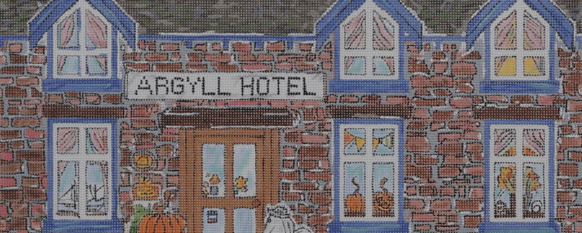 A close-up of a cross-stitch or needlepoint textile design depicting the front facade of the Argyll Hotel, featuring a brick wall, blue-trimmed windows, a central door, and embroidered autumn details.